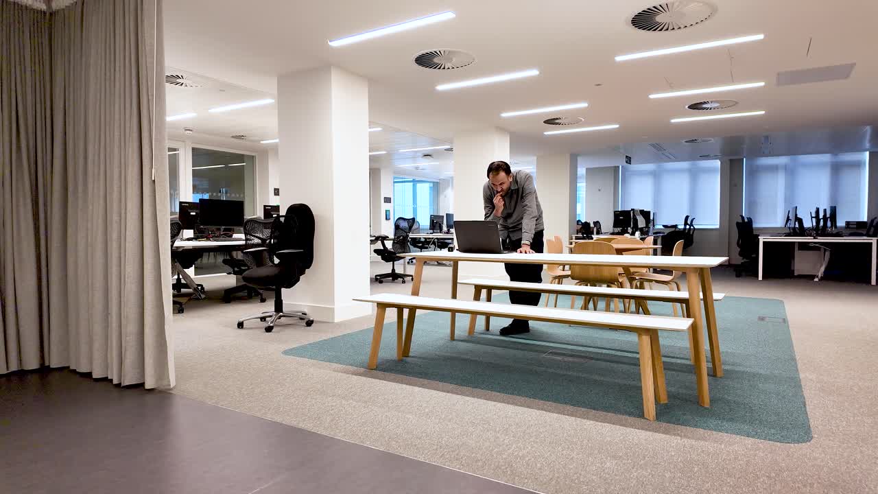 Man working with laptop while leaning over a desk in a large, empty contemporary office