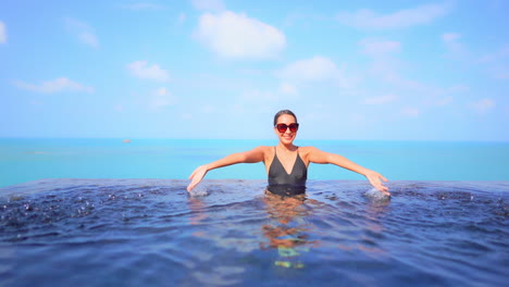 With-the-ocean-as-a-backdrop,-a-pretty-young-woman-playfully-splashes-water-in-the-swimming-pool