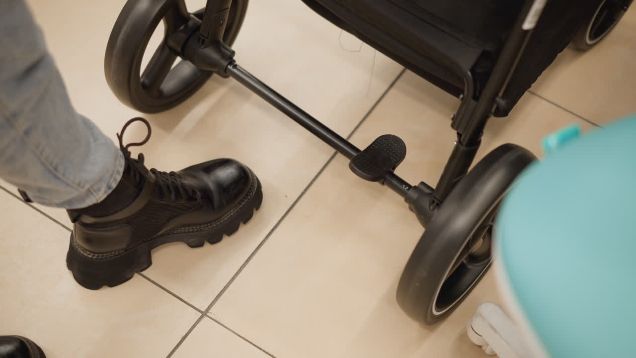 Leg view of young woman in black boots and denim trousers examining stroller wheel roller functionality on tile floor in brightly lit baby gear department demonstration of ergonomic design features