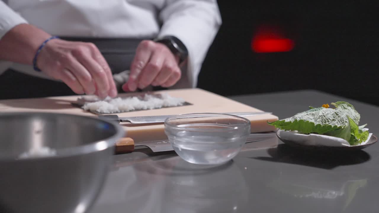A static shot captures a sushi chef carefully spreading rice onto seaweed, preparing to make sushi rolls. Perfect for culinary, sushi preparation, and professional kitchen content.