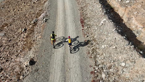 Fantástica-Toma-Aérea-De-Dos-Hombres-En-Sus-Bicicletas-De-Montaña-Parlantes-Recorriendo-La-Carretera-En-Un-Paisaje-Desértico