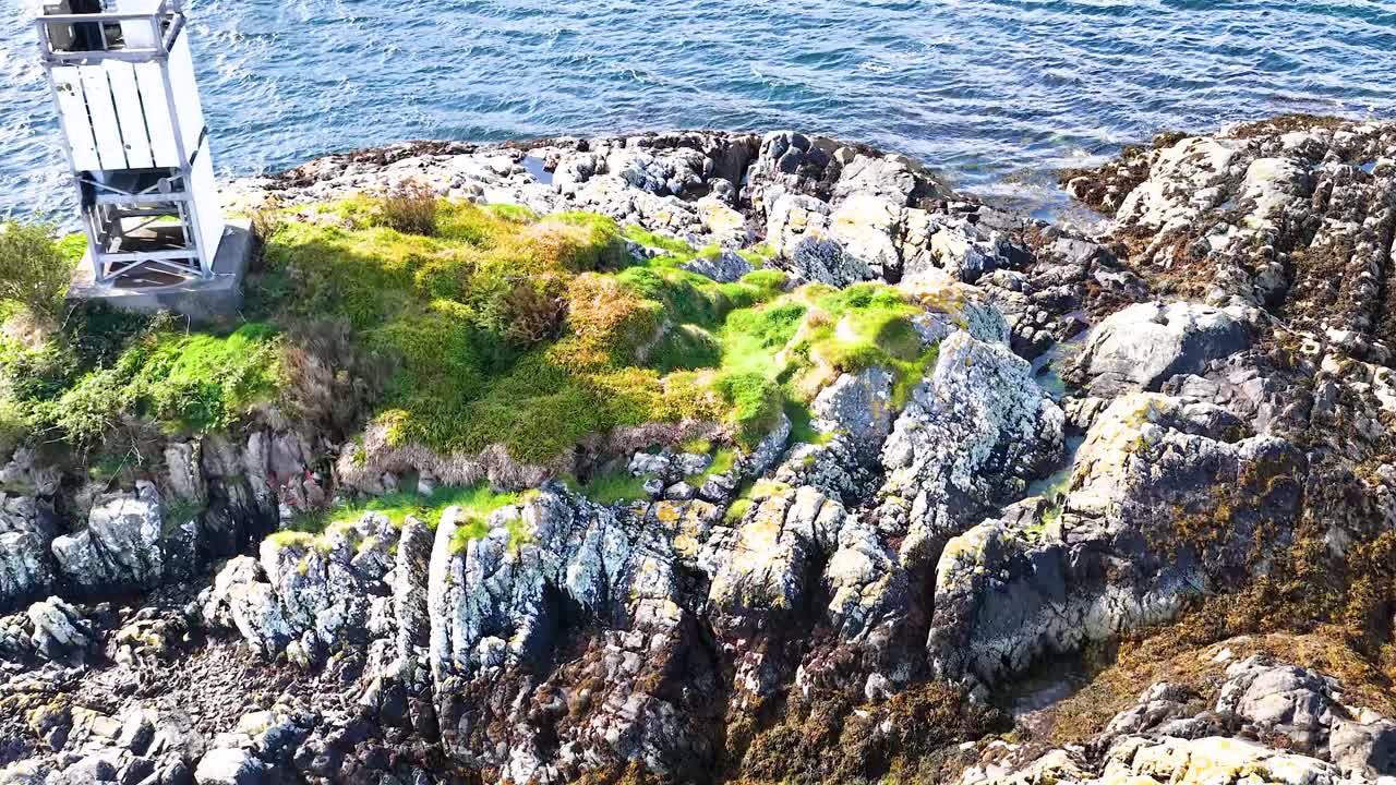 Drone glides over rocky island with lighthouse, lush greenery, and clear coastal waters in sunlight