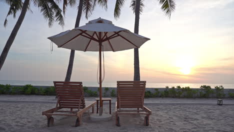 Two-empty-beach-loungers-under-a-sun-umbrella-on-a-sandy-beach-looking-out-on-a-colorful-setting-sun