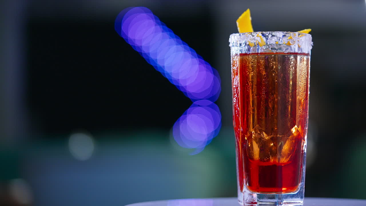 Misted long glass with red drink. Rotating table with cocktail decorated with orange peel. Close up. Blurred backdrop.