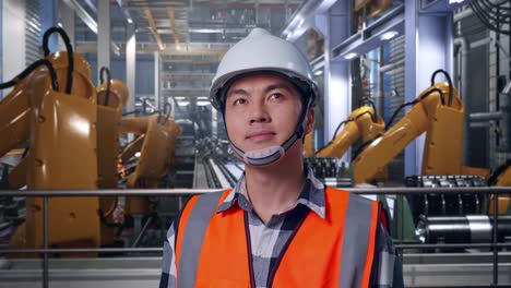 close up of asian male engineer with safety helmet looking around while standing with industrial robotic arms working