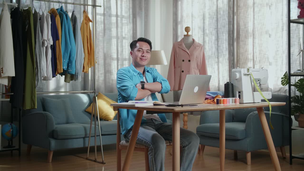 Asian Male Designer Working On A Laptop And Smiling To The Camera With Arms Crossed While Designing Clothes In The Studio