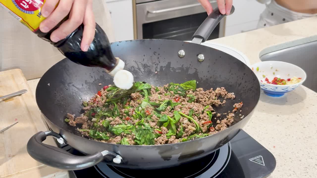 A person prepares Thai stir fry pork with basil in a hot wok, adding fish sauce and seasonings in a bright, modern kitchen setting