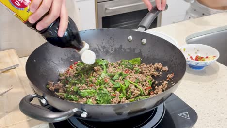 a person prepares thai stir fry pork with basil in a hot wok, adding fish sauce and seasonings in a bright, modern kitchen setting