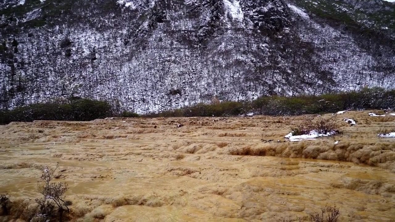 Uma ampla e lenta panorâmica revelando um vasto campo de terraços de tufo mineral amarelo-dourado que se estendem em direção a uma encosta íngreme coberta por árvores salpicadas de neve.