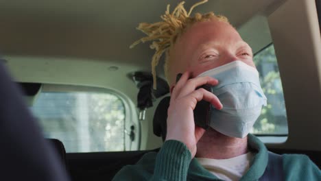 feliz albino hombre afroamericano con máscara de cara sentado en el coche hablando en el teléfono inteligente