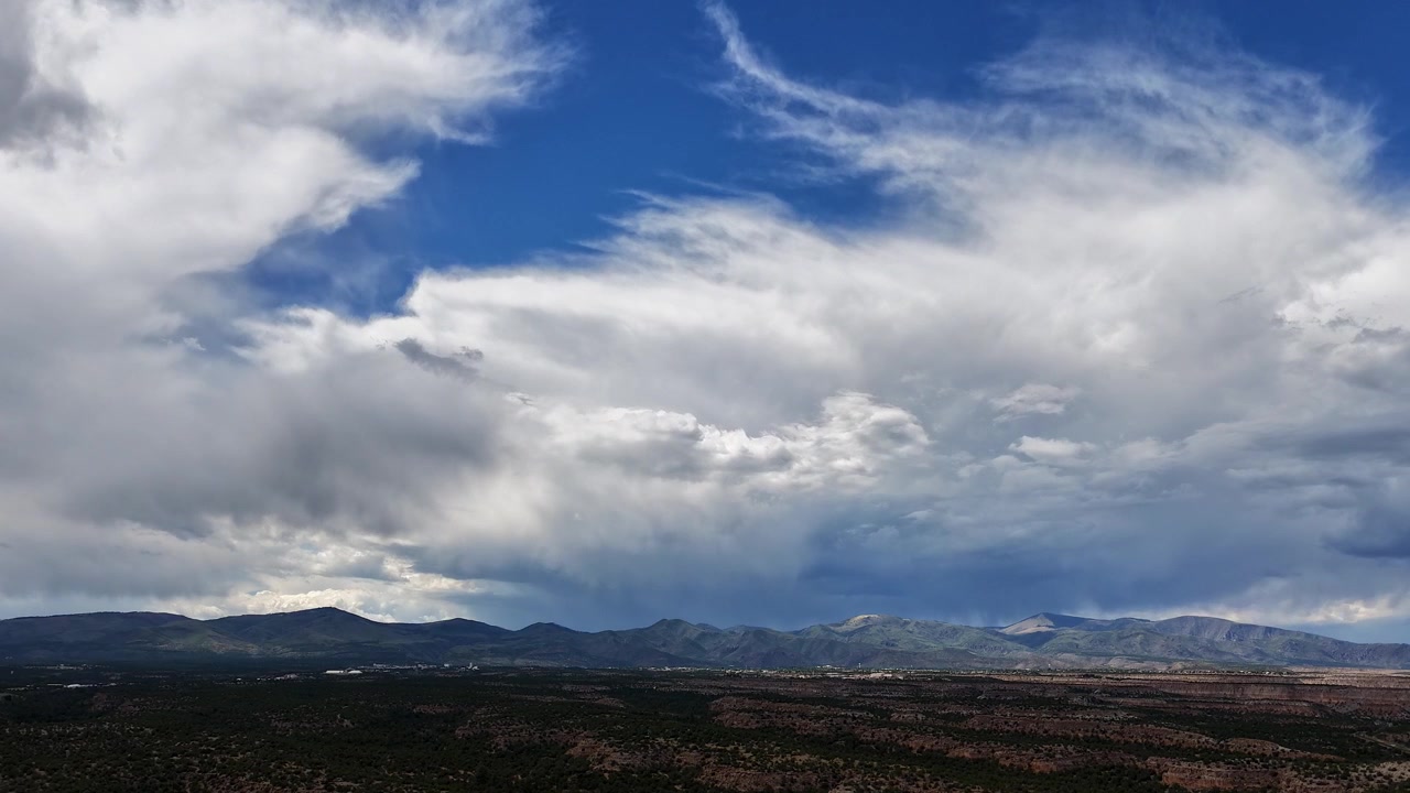 Time-lapse 4K da drone che cattura nuvole nebbiose che si muovono rapidamente sopra le vaste mesas e le Jemez Mountains. Ombre dinamiche cadono sul fondovalle mentre il tempo cambia sul paesaggio arido