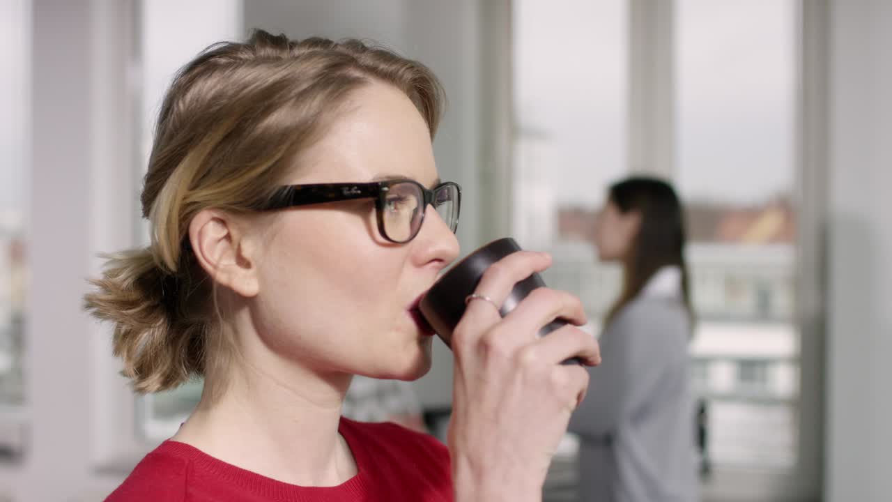 누군가의 말을 듣는 동안 컵을 마시는 안경을 가진 자신감 있는 갈색 머리 여자