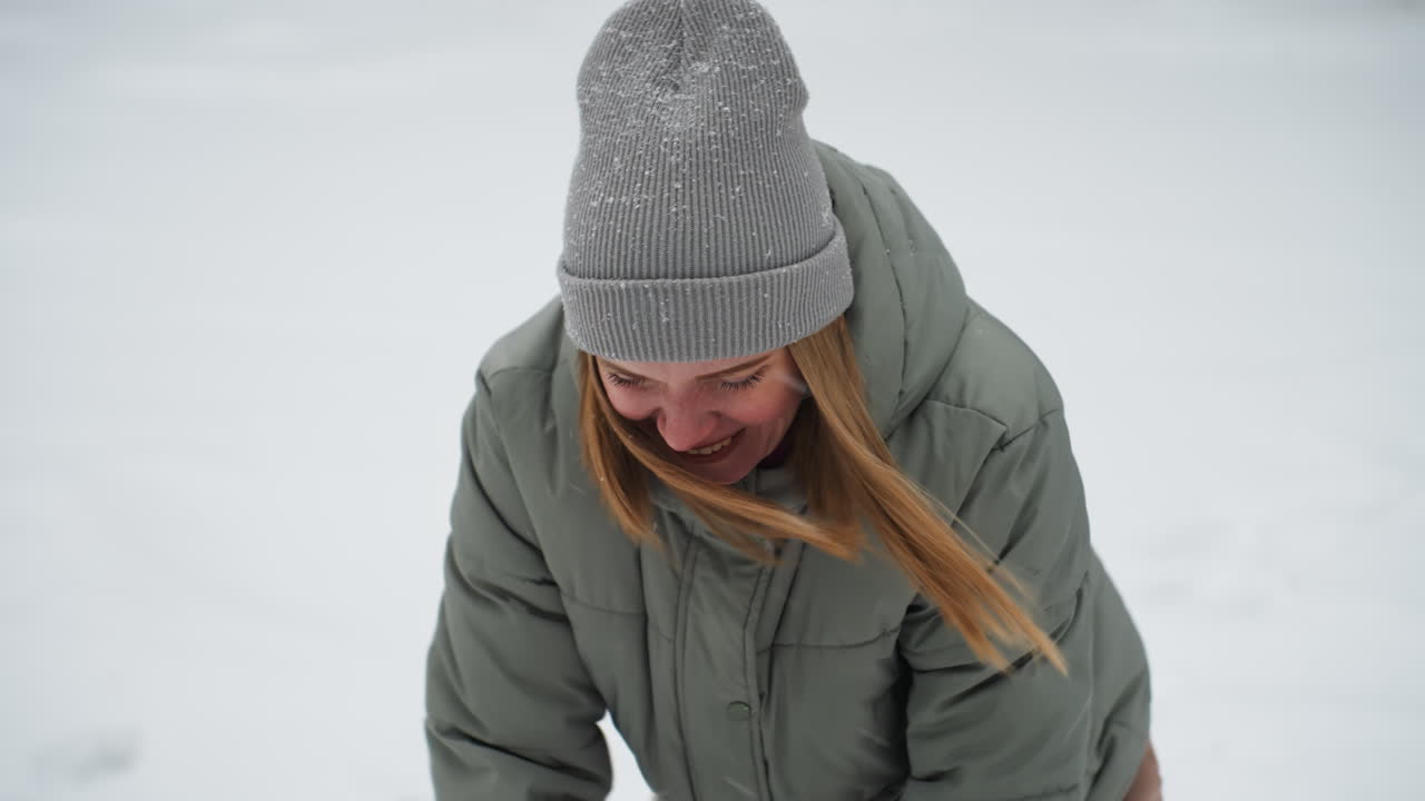 Young woman in green puffer jacket and gray beanie leans forward while laughing joyfully during snowy winter day, face partially visible, snowflakes sprinkled on hat, creating candid and heartfelt moment