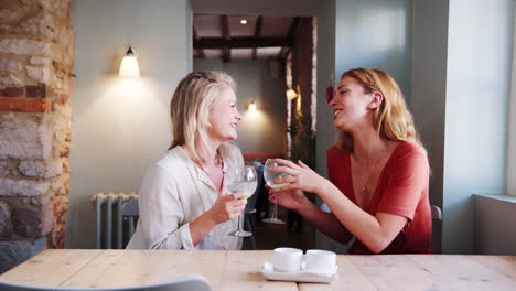 two young adult blonde women holding wine glasses talking with each other sitting at a table in a pub, close up, side view