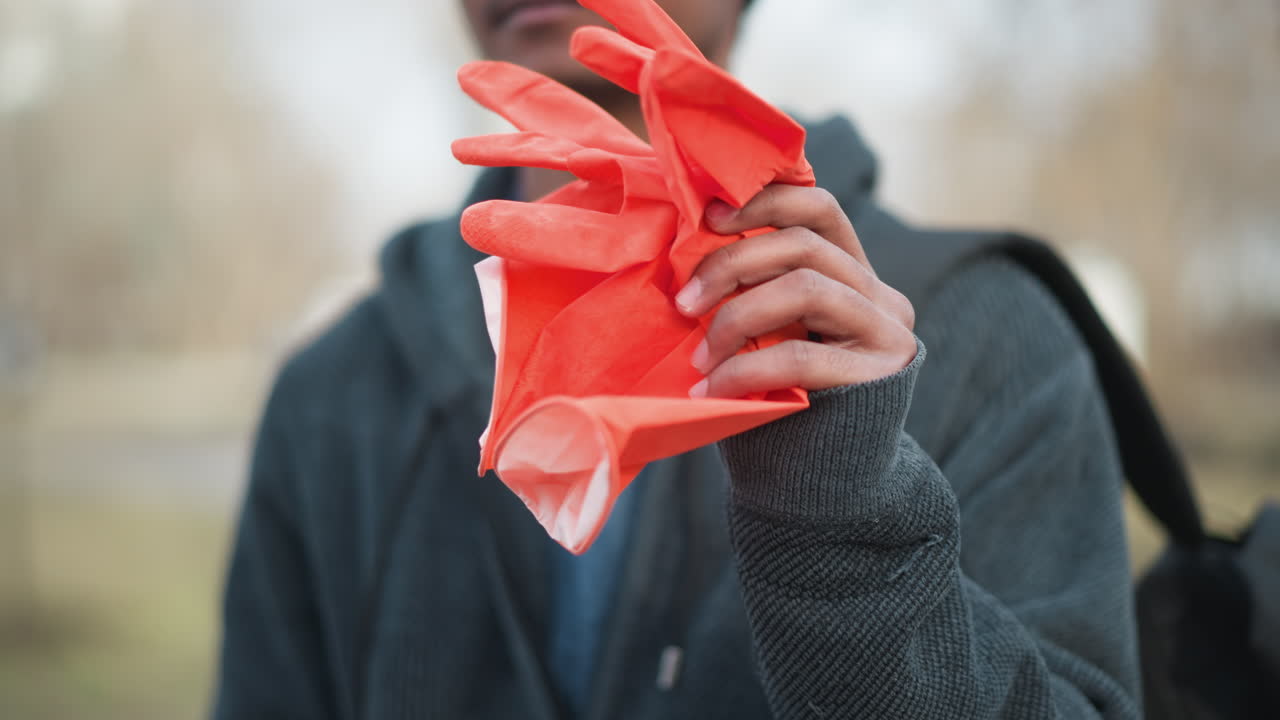 Close-up of person holding bright orange rubber gloves outdoors, wearing dark textured hoodie and backpack strap, symbolizing readiness for cleaning, hygiene, volunteer service