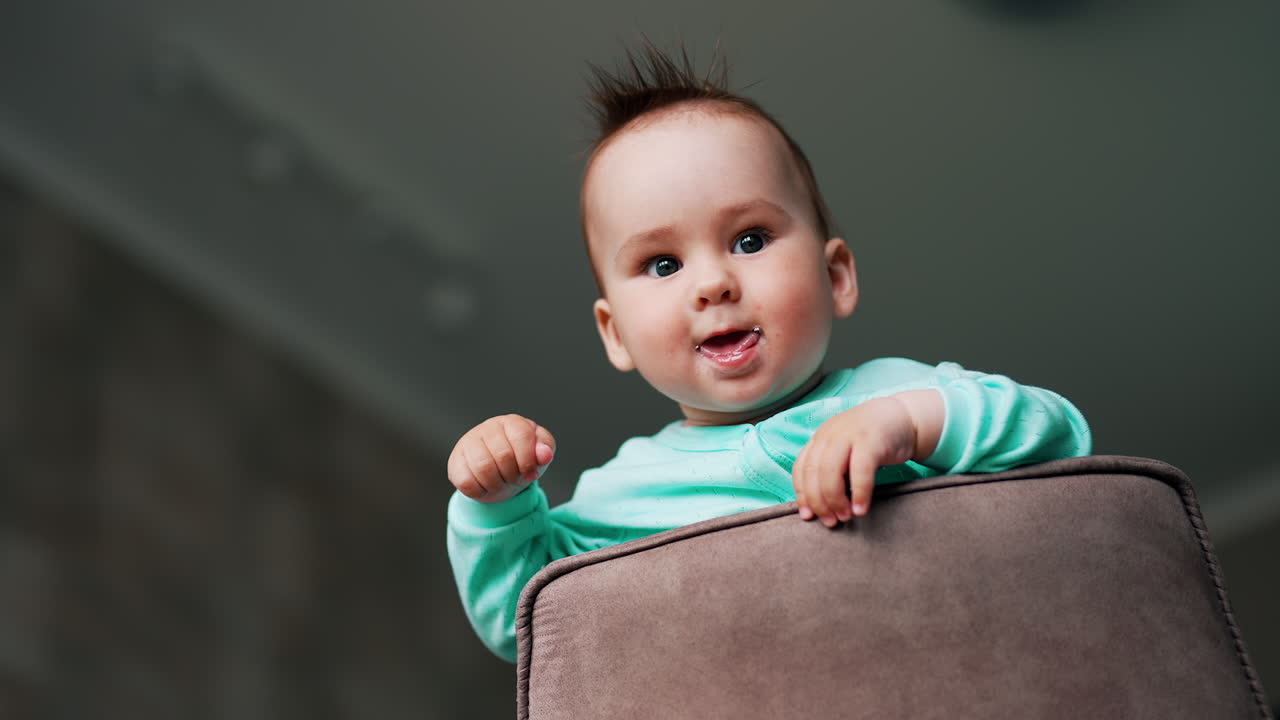 Little adorable baby stands on the chair looking down at camera. Nice Caucasian kid showing tongue and waving hand. Low angle view.