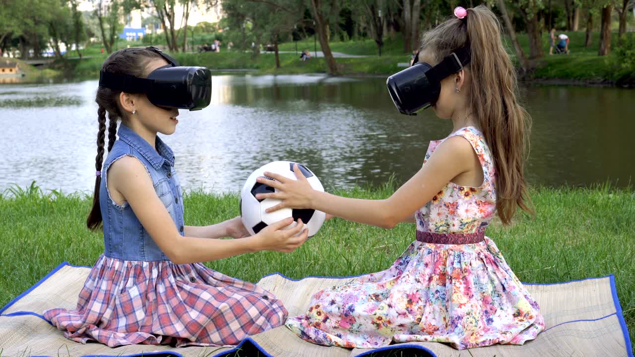 dos niñas con gafas de realidad virtual están sentadas una frente a otra en el parque en la hierba al atardecer, con una pelota de fútbol en las manos y girándola.