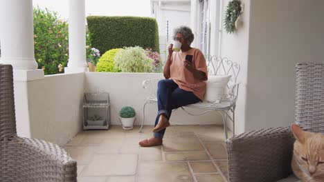 mujer afroamericana mayor que usa un teléfono inteligente mientras bebe café sentada en el porche de la casa