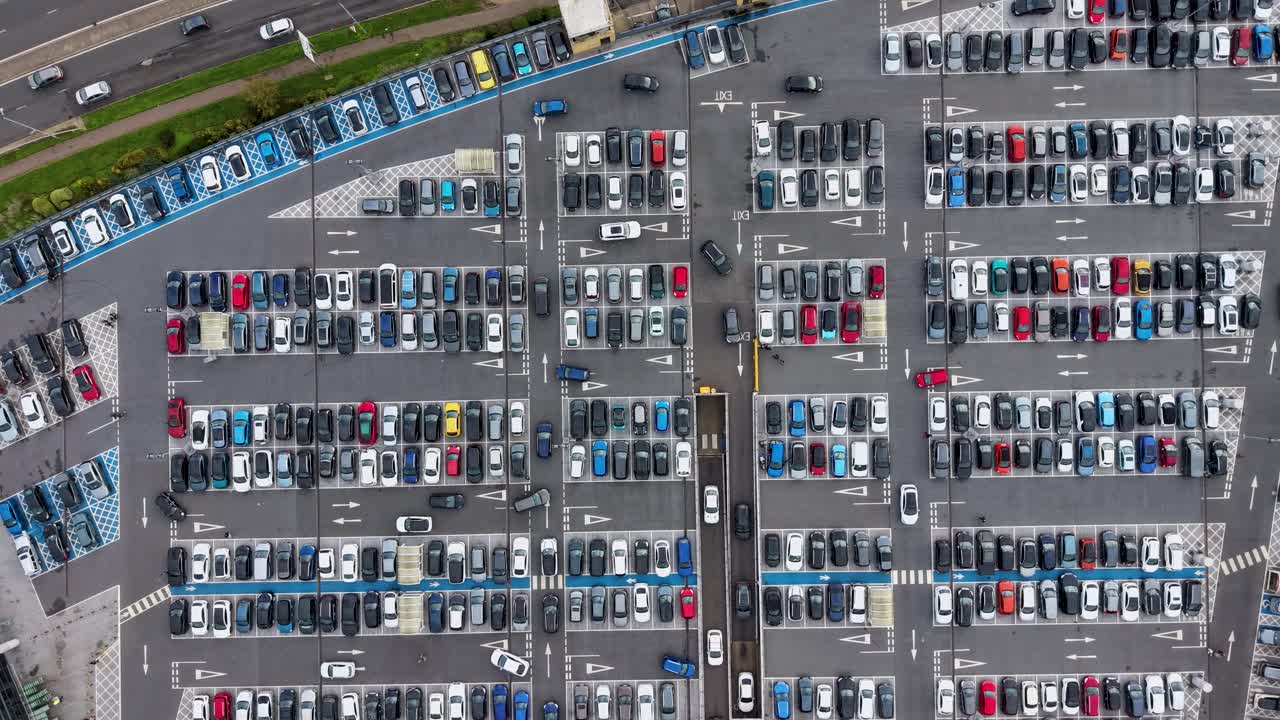 Top-down drone timelapse of Water Garden Harlow, UK, car park. Cars drive around the busy car park, with lower level visible, and traffic flow on main road at top, capturing vibrant urban movement