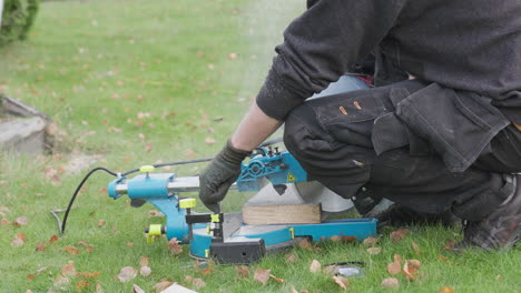 A close-up of a worker operating a blue miter saw on a grassy lawn with scattered leaves.