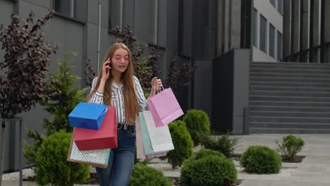 positive young girl with bags talking on mobile phone about sales in shopping mall in black friday