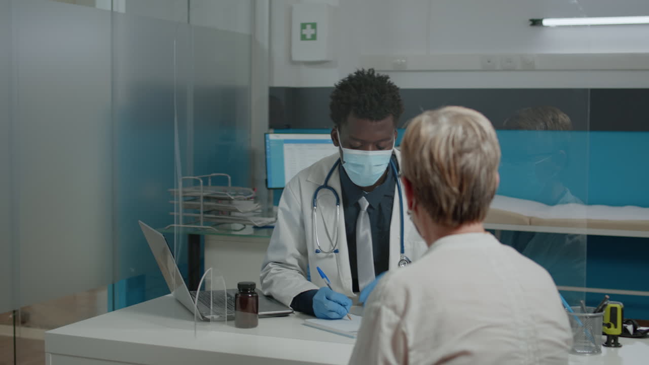 Medic with white coat sitting at desk while talking to elder woman