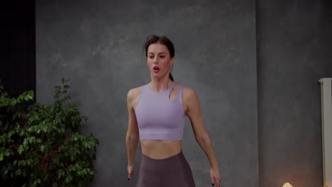 chica bruneta atlética confiada en top púrpura haciendo sentadillas y saltando en un apartamento moderno mientras hace deportes en casa. chica brune confiada haciendo deportes en casa en un departamento moderno con paredes grises