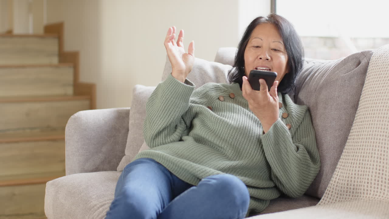 Senior Asian woman relaxing on couch, chatting on smartphone, enjoying cozy home moment