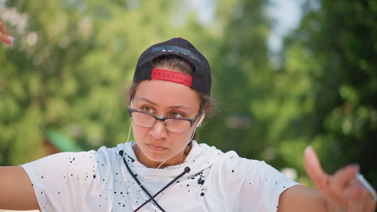 공원에서 클로즈업으로 촬영한 생동감 넘치는 상반신 댄스 동작, 뒤로 쓴 모자와 안경을 착용하고 이어폰을 낀 채 안무를 연습하는 젊은 공연자, 활기찬 손동작, 자연스러운 리허설 장면, 여름 햇살