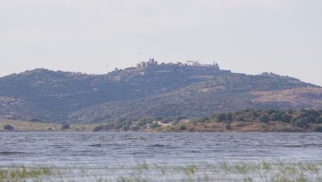 A-serene-ground-level-view-of-Monsaraz-as-seen-from-the-lakeside,-highlighting-the-tranquil-waters-and-the-picturesque-medieval-village-perched-atop-the-hill-in-the-background