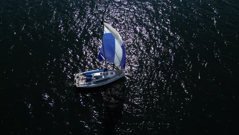 Aerial-view-of-a-sailboat-sailing-on-the-dark-water-of-the-baltic-sea,-slow-orbital-drone-shot,-high-contrast-scene,-blue-sail