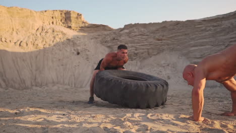 dos atletas entrenan en modo activo en la playa haciendo flexiones y empujando una gran rueda