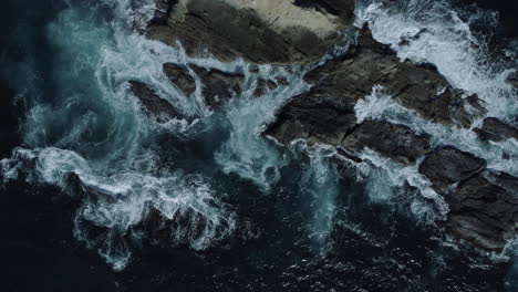 aerial shot from low to high of blue ocean water crashing on rocks