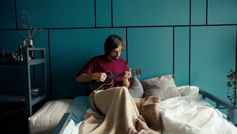 Ein-Brünetter-Mann-In-Einem-Dunkelroten-Hemd-Liegt-Auf-Einem-Sofa,-In-Eine-Decke-Gehüllt,-Und-Spielt-Gitarre-Vor-Einer-Türkisfarbenen-Wand