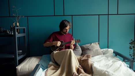 A-brunette-man-in-a-dark-red-shirt-lies-on-a-sofa-wrapped-in-a-blanket-and-plays-the-guitar-against-a-turquoise-wall