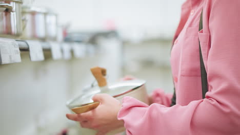 hand of female shopper inspecting a modern cookware pot in a kitchenware store, the shopper is carefully lifting the pot's lid with a wooden handle, surrounded by other displayed pots in the store