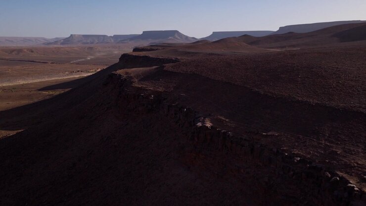 Una telecamera aerea si muove lungo scarpate desertiche rocciose, rivelando ripidi pendii in ombra, ampi altipiani e distanti mesa dalla cima piatta in Marocco