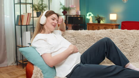 Pregnant woman eyes closed listens calm music with earbuds strokes belly peaceful indoor rest relax