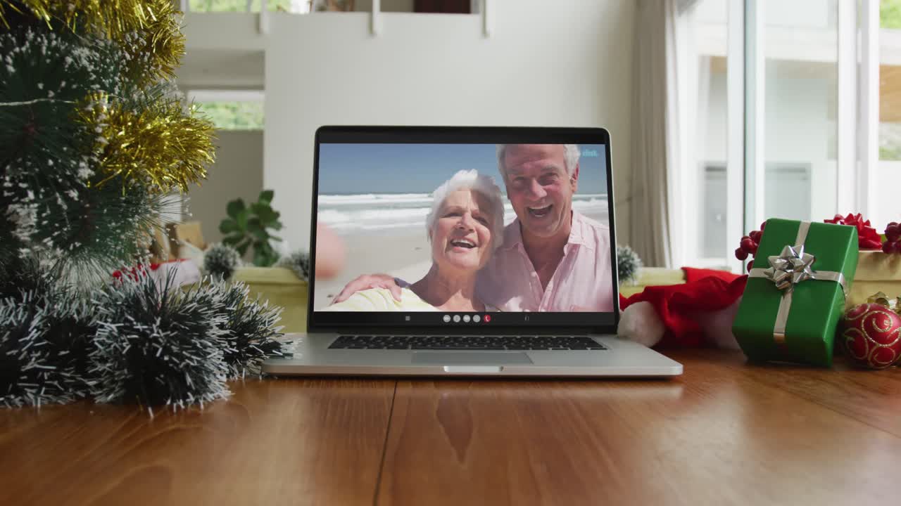 sonriente pareja caucásica en la playa en navidad llamada de video en computadora portátil