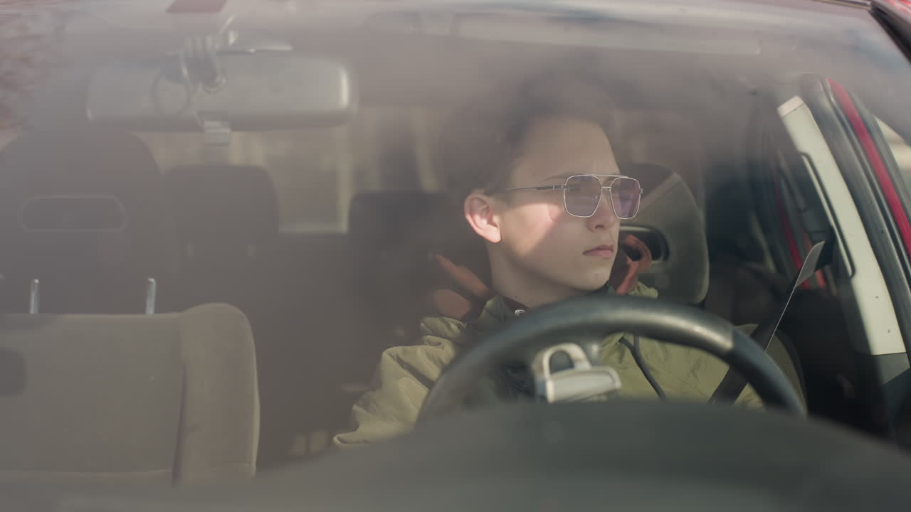 young boy in parked car seen through windshield wearing green winter jacket and glasses while removing seatbelt and holding steering wheel with one hand during daylight in outdoor setting