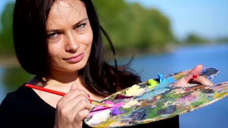 summer,outdoors, portrait of a beautiful forty years old brunette woman artist, close-up of a palette with paints, the woman artist mixes paints with a brush on the palette