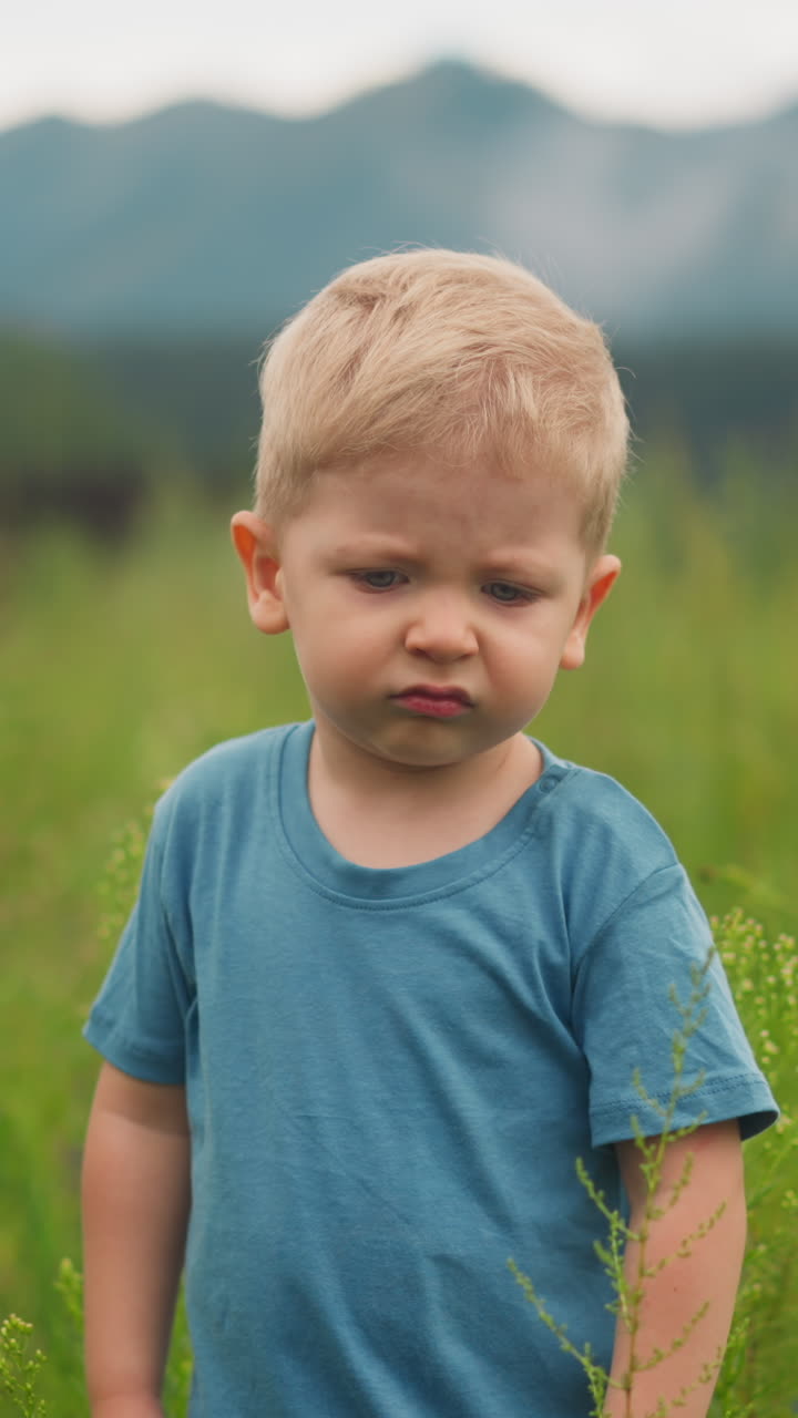 Unhappy little boy in blue t-shirt grimaces walking along meadow with high green grass in valley on cloudy nasty day closeup slow motion