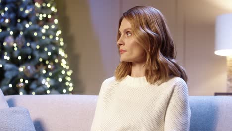 close up portrait of joyful young female sitting in cozy room with glowing tree in good mood, looking away and smiling to camera