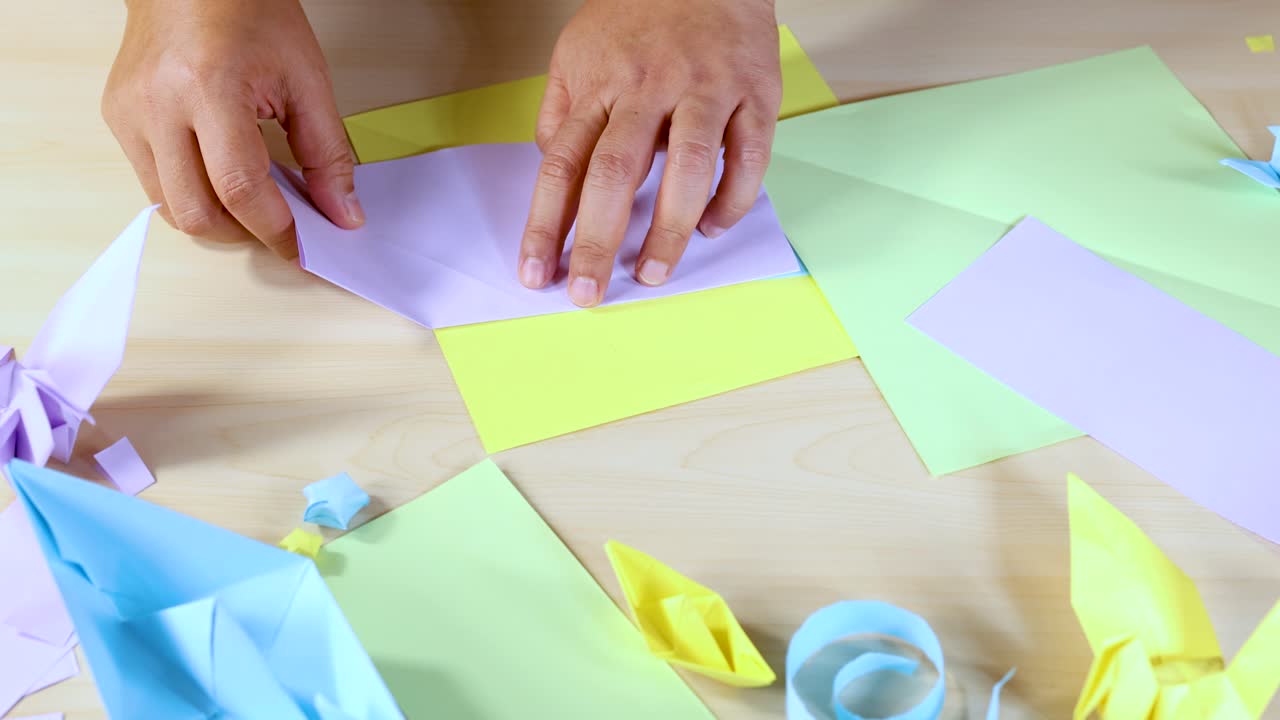 Person folding pastel origami paper at desk, surrounded by colorful finished models, overhead view