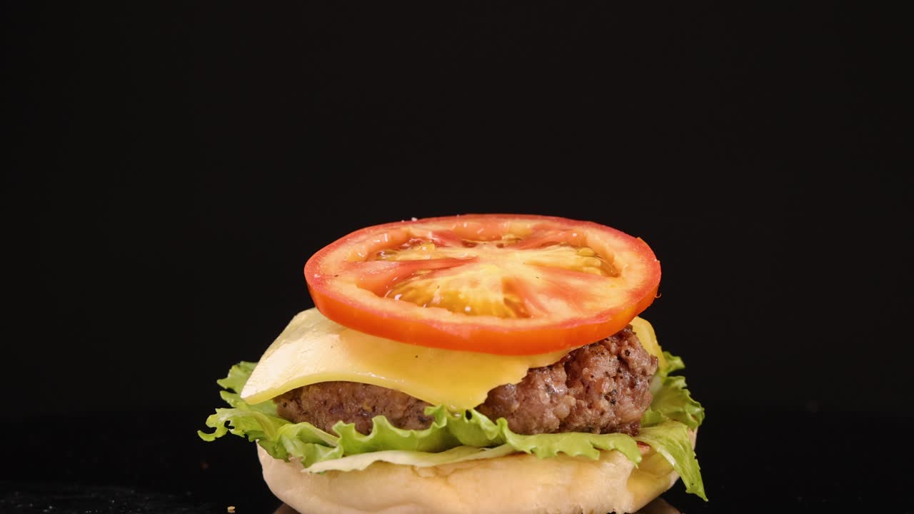 A hand delicately lifts a tomato slice from a cheeseburger with lettuce, cheese, and onion against a black background. Studio lighting, static camera