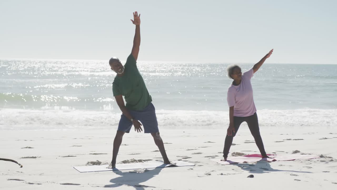 Happy senior african american couple doing yoga and stretching at beach, in slow motion