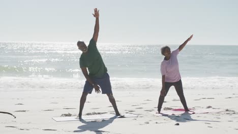 Happy-senior-african-american-couple-doing-yoga-and-stretching-at-beach,-in-slow-motion