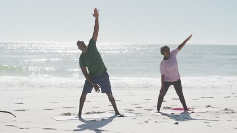 happy senior african american couple doing yoga and stretching at beach, in slow motion