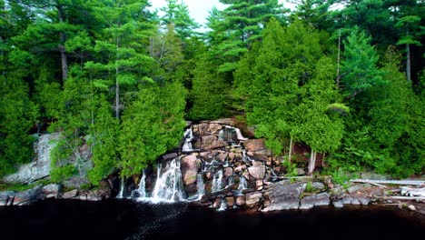 Water-cascading-down-rocky-waterfall-between-lush-green-forest-into-a-dark-colored-lake-view-from-above