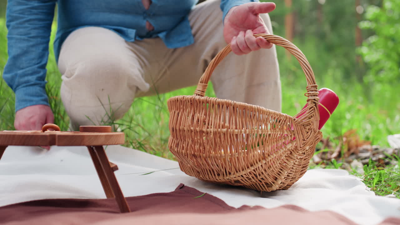 푸른색 셔츠와 베이지색 바지를 입은 남성이 허리를 굽혀 작은 테이블이 놓인 매트 위에 피크닉 바구니를 내려놓는 모습이에요. 초록색 잔디와 햇빛으로 둘러싸인 평화로운 야외 피크닉 장면을 낮은 뷰로 담았습니다.